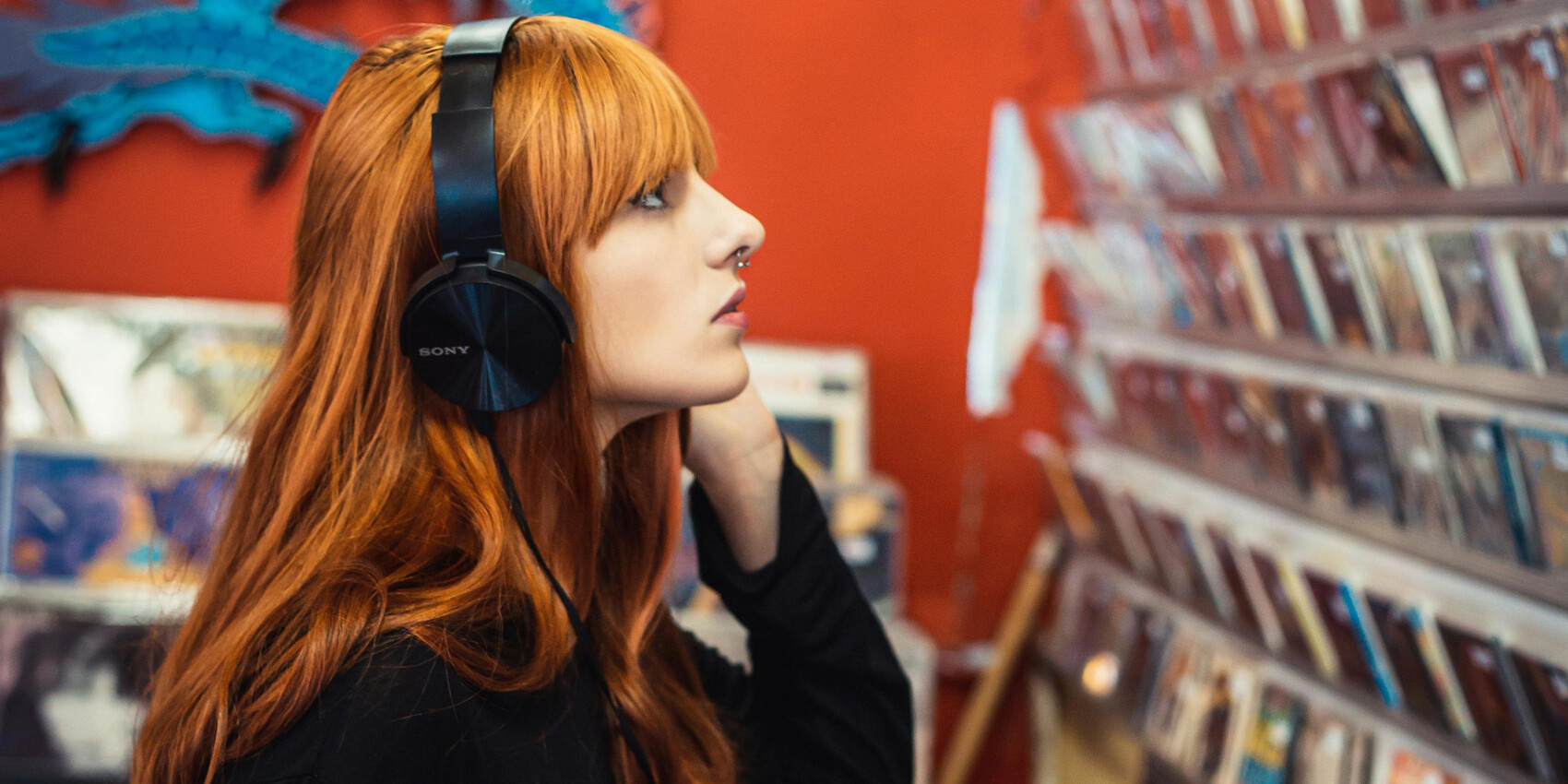 A person with long red hair wearing headphones stands in a music store, looking at a wall display of CDs. The background is red with a blue decorative element visible—it's the perfect place to discover music for less than a song.