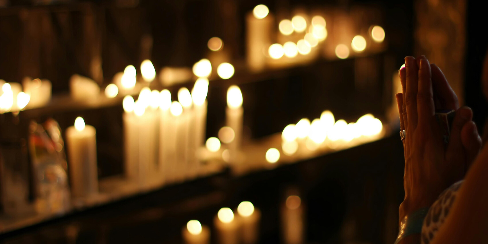 A person with hands clasped in prayer faces a shelf filled with numerous lit candles, creating a warm, soft glow in a dimly lit setting—a quiet scene that prompts reflection on how My Canada doesn't include religiosity.