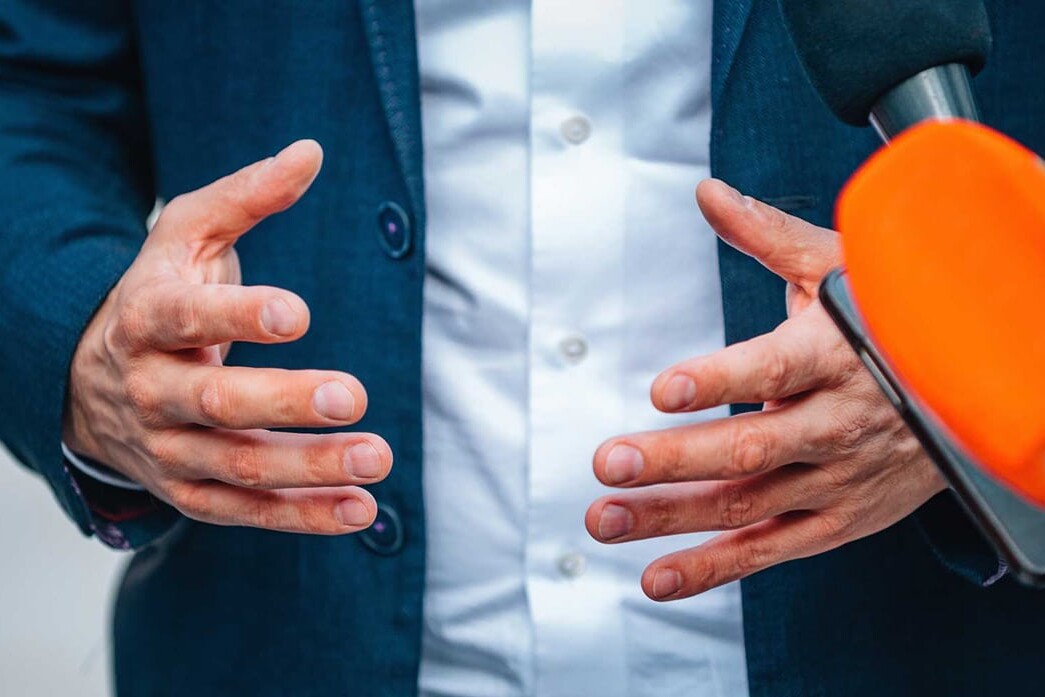 John Tory, well beyond the bland, gestures with his hands in a blue suit while speaking to the media, surrounded by microphones, including one with an orange foam cover.
