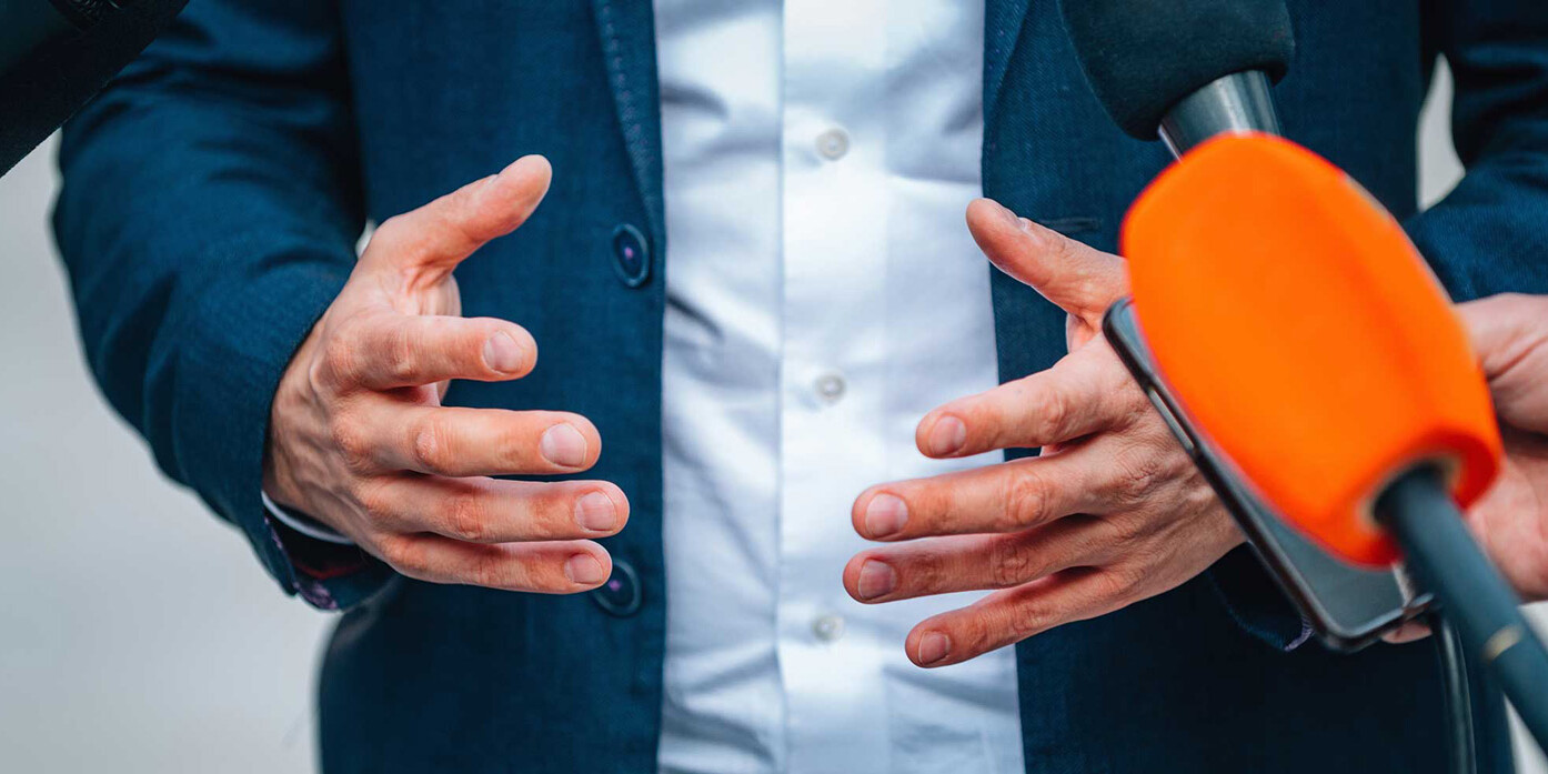 John Tory, well beyond the bland, gestures with his hands in a blue suit while speaking to the media, surrounded by microphones, including one with an orange foam cover.