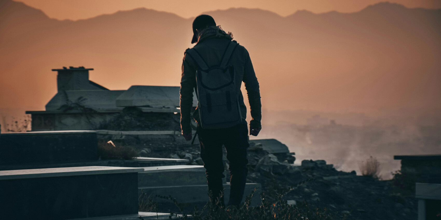 A person wearing a backpack walks among gravestones at sunset, with mountains and an orange sky in the background—a somber, silhouetted scene that echoes the solitude found in stories from the 2007 Survey of Afghans.
