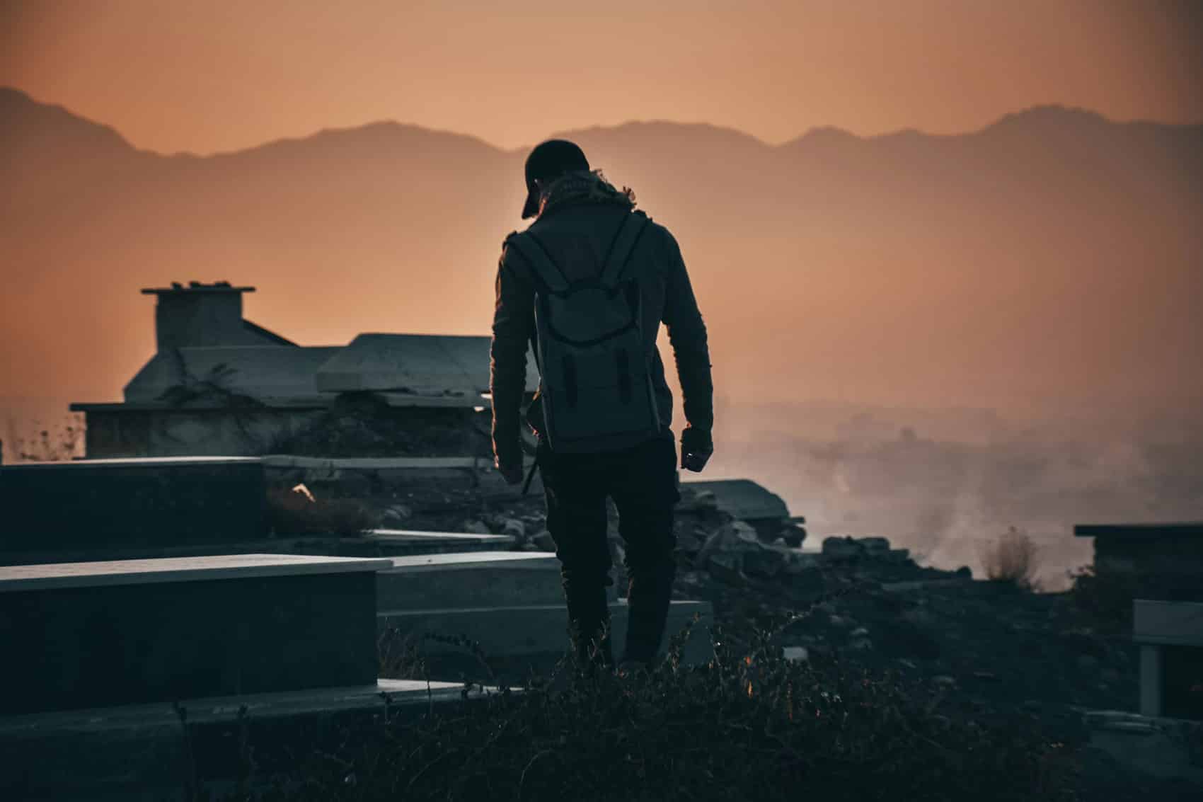 A person wearing a backpack walks among gravestones at sunset, with mountains and an orange sky in the background—a somber, silhouetted scene that echoes the solitude found in stories from the 2007 Survey of Afghans.