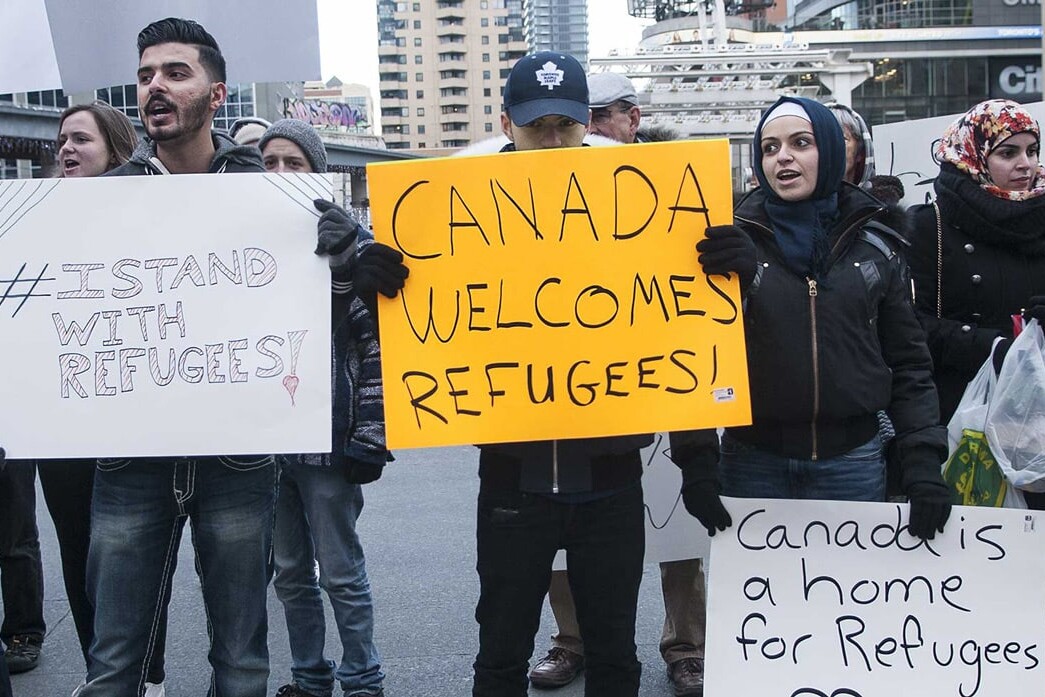 A diverse group of people hold signs supporting refugees at an outdoor rally in a city. Signs read “#Stand With Refugees,” “Canada Welcomes Refugees!” and “We’re no bigots—Canada is a home for refugees.”.