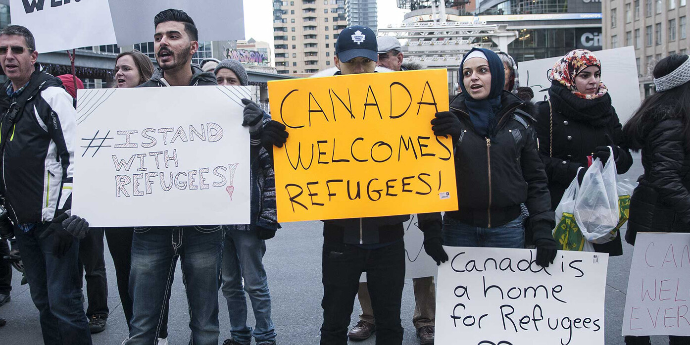 A diverse group of people hold signs supporting refugees at an outdoor rally in a city. Signs read “#Stand With Refugees,” “Canada Welcomes Refugees!” and “We’re no bigots—Canada is a home for refugees.”.