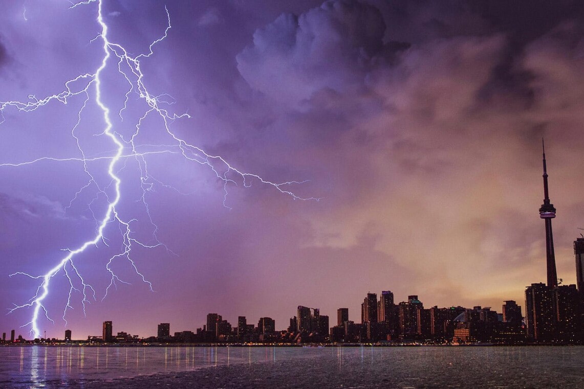 A dramatic lightning bolt strikes the sky above a city skyline at dusk, tall buildings and a prominent tower silhouetted against purple and orange clouds, reflected in calm water below—much like the question: Canadians are ready for a carbon tax. Is anyone listening?.