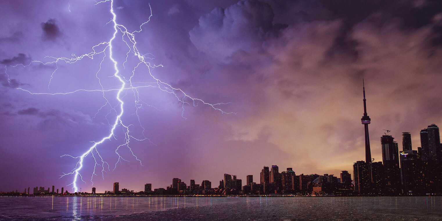 A dramatic lightning bolt strikes the sky above a city skyline at dusk, tall buildings and a prominent tower silhouetted against purple and orange clouds, reflected in calm water below—much like the question: Canadians are ready for a carbon tax. Is anyone listening?.