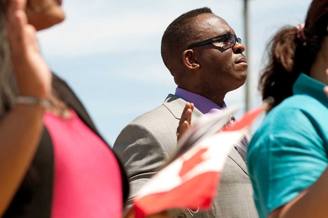 A group of people, possibly immigrants, stand outdoors raising their right hands during a ceremony. One person holds a Canadian flag under the clear, sunny sky. Are immigrants natural conservatives? The scene reflects diversity and unity.