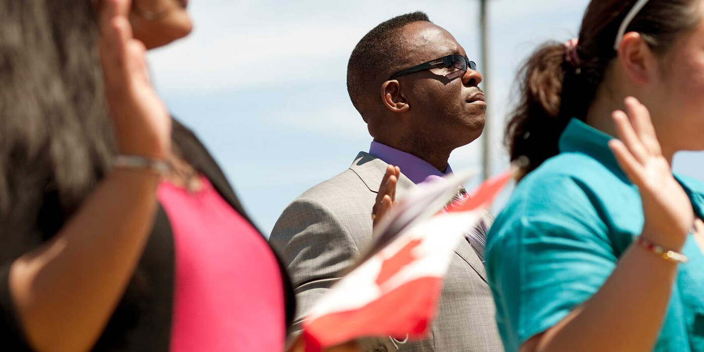 A group of people, possibly immigrants, stand outdoors raising their right hands during a ceremony. One person holds a Canadian flag under the clear, sunny sky. Are immigrants natural conservatives? The scene reflects diversity and unity.
