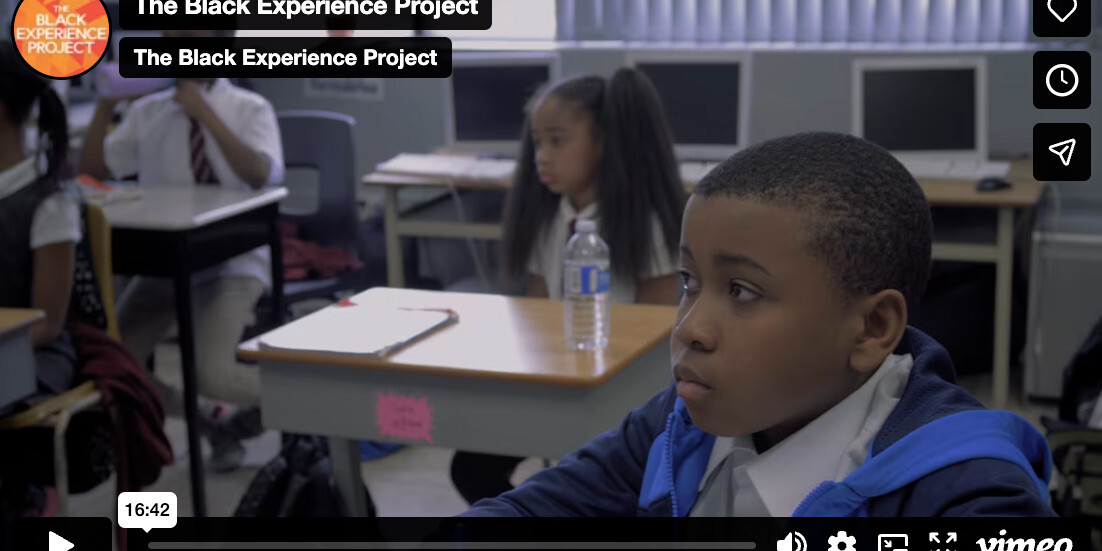 A young boy sits at a desk in a classroom, appearing focused, as The Black Experience Project is displayed on the screen. Other students are seated in the background, creating an atmosphere of learning and reflection.