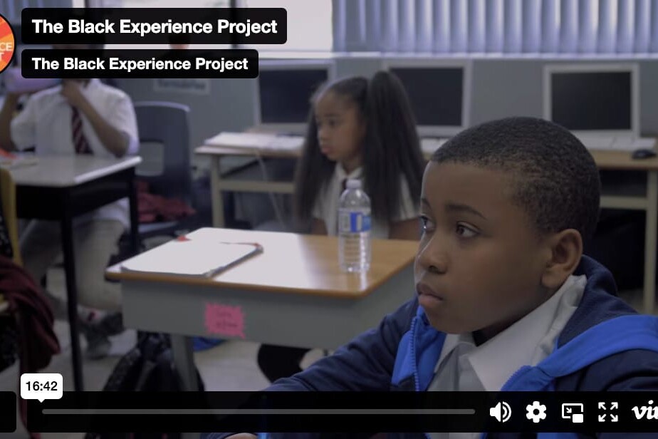 A young boy sits at a desk in a classroom, appearing focused, as The Black Experience Project is displayed on the screen. Other students are seated in the background, creating an atmosphere of learning and reflection.