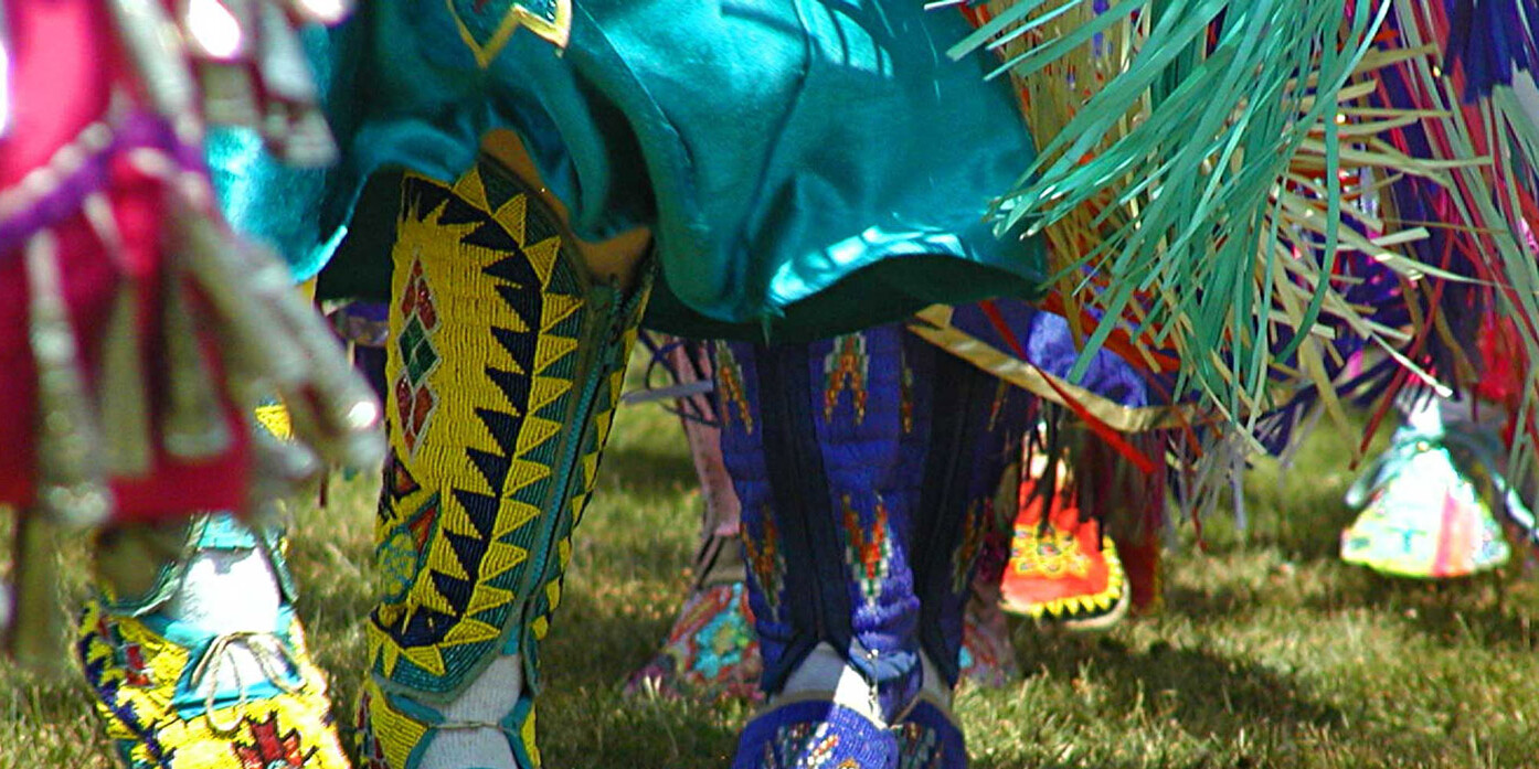 Close-up of dancers’ feet and legs in colorful, beaded moccasins and fringed regalia on grass at a powwow—showcasing what success might look like for young Aboriginals embracing tradition and cultural pride.