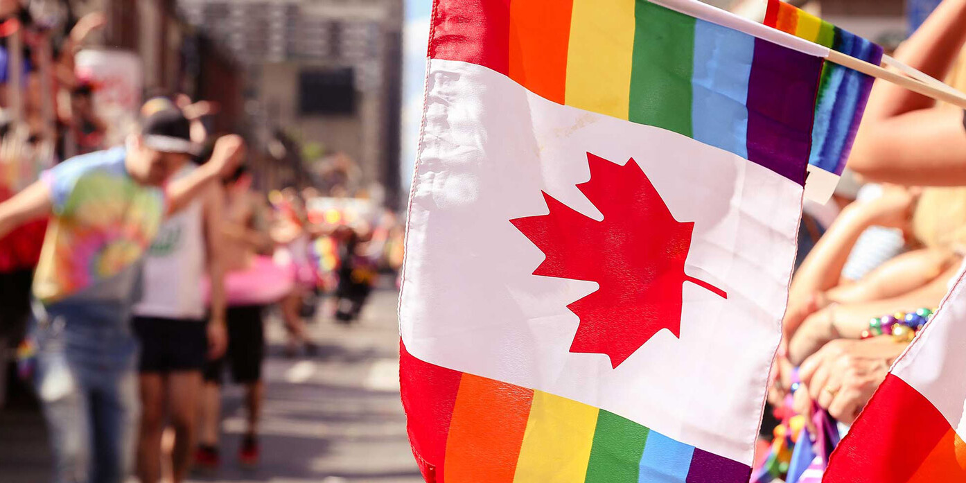 Two Canadian flags with rainbow stripes are held at a vibrant outdoor parade. People in colorful clothing and rainbow accessories line the street, celebrating diversity and inclusion—a counterpoint to claims that Harper has not shifted Canadians' opinions.