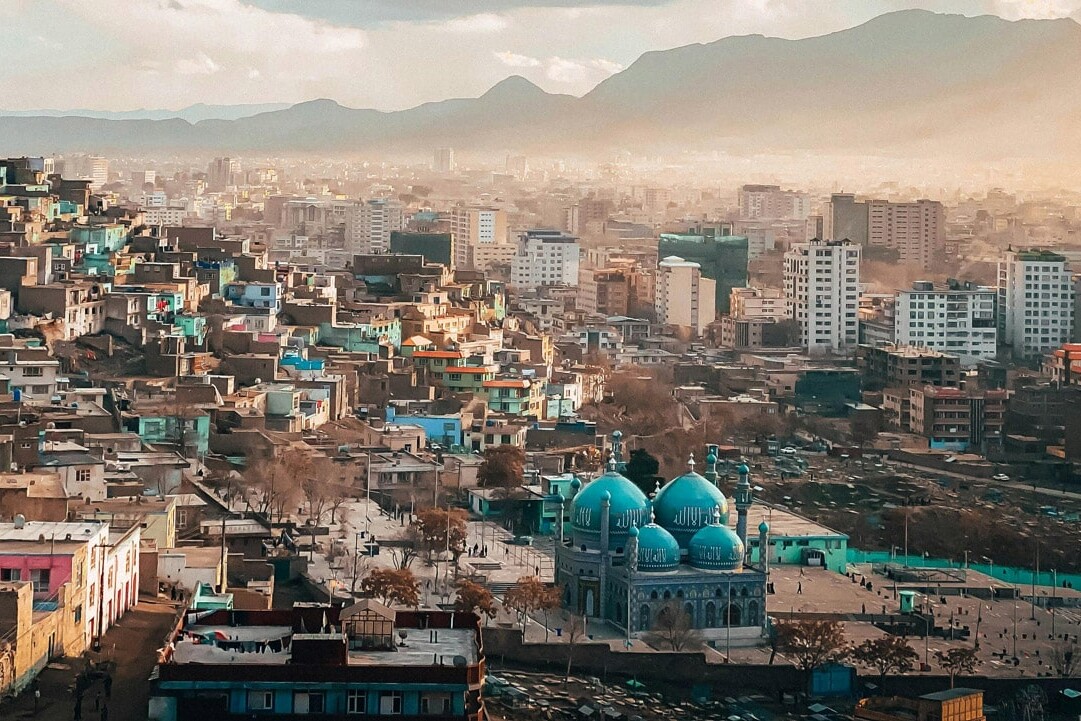A sprawling cityscape with colorful hillside houses, a blue-domed mosque at its center, and modern buildings rising in the background, set against distant mountains under a partly cloudy sky. We support the war (for now).