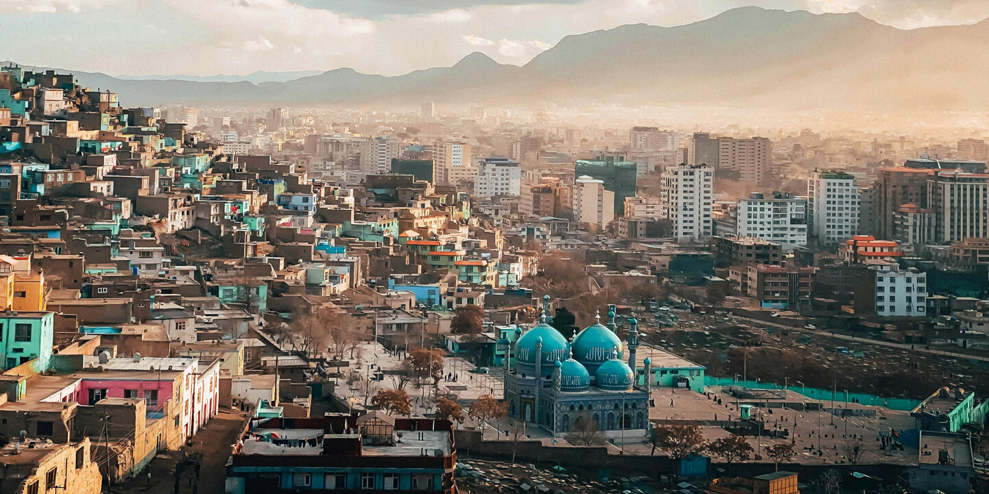 A sprawling cityscape with colorful hillside houses, a blue-domed mosque at its center, and modern buildings rising in the background, set against distant mountains under a partly cloudy sky. We support the war (for now).