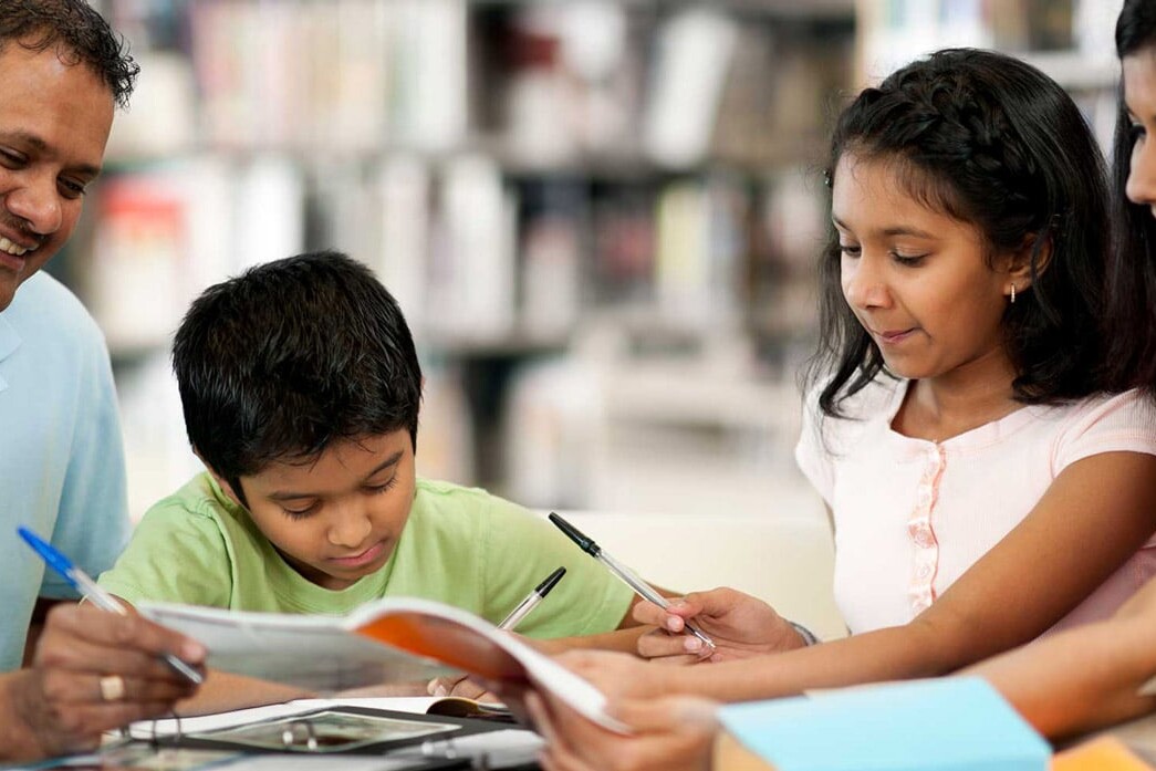A smiling family of four, including two adults and two children, sit together at a table in a library, reading books and writing—perhaps even exploring topics like why politicians fixate on the ethnic vote—with shelves of books in the background.