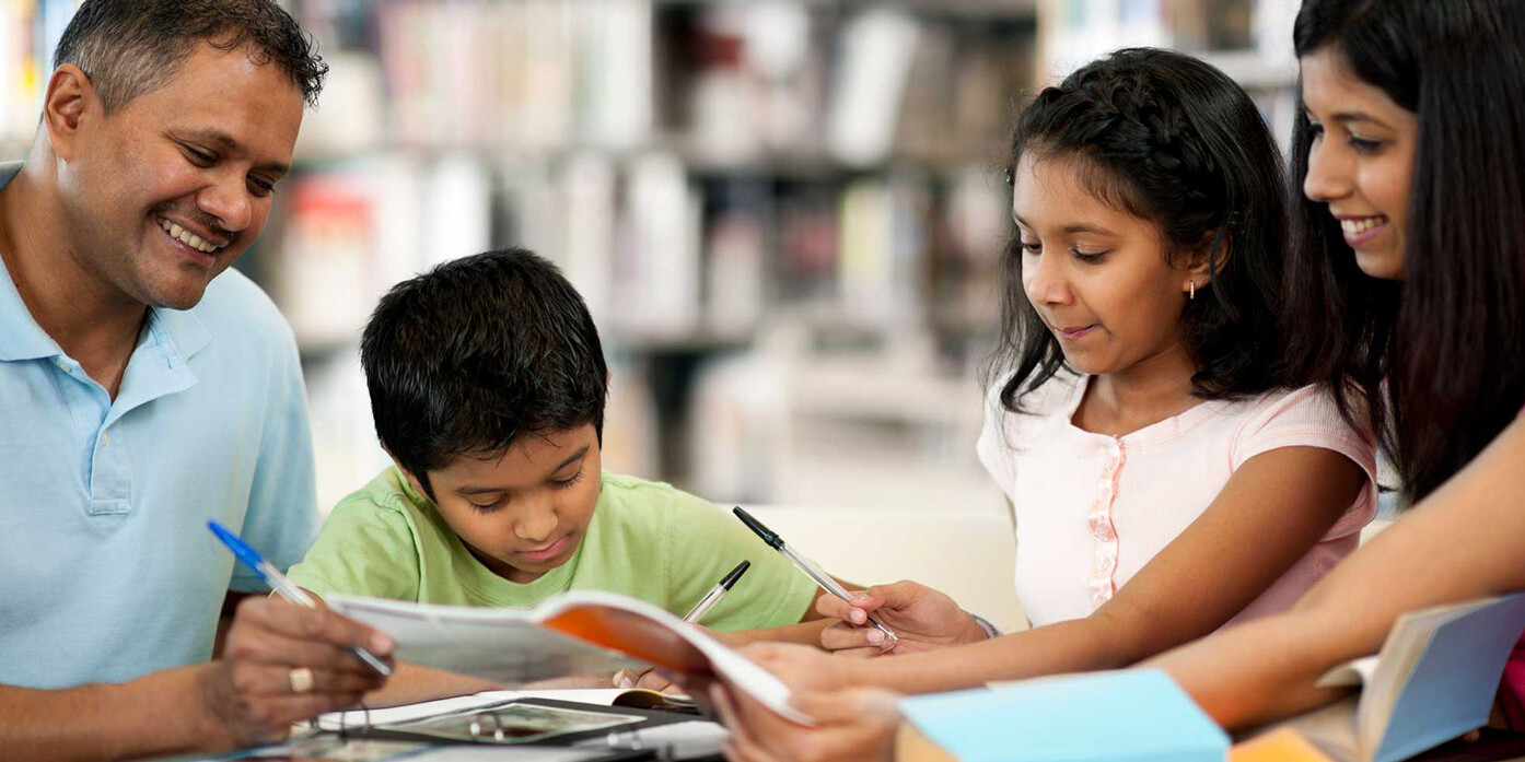 A smiling family of four, including two adults and two children, sit together at a table in a library, reading books and writing—perhaps even exploring topics like why politicians fixate on the ethnic vote—with shelves of books in the background.