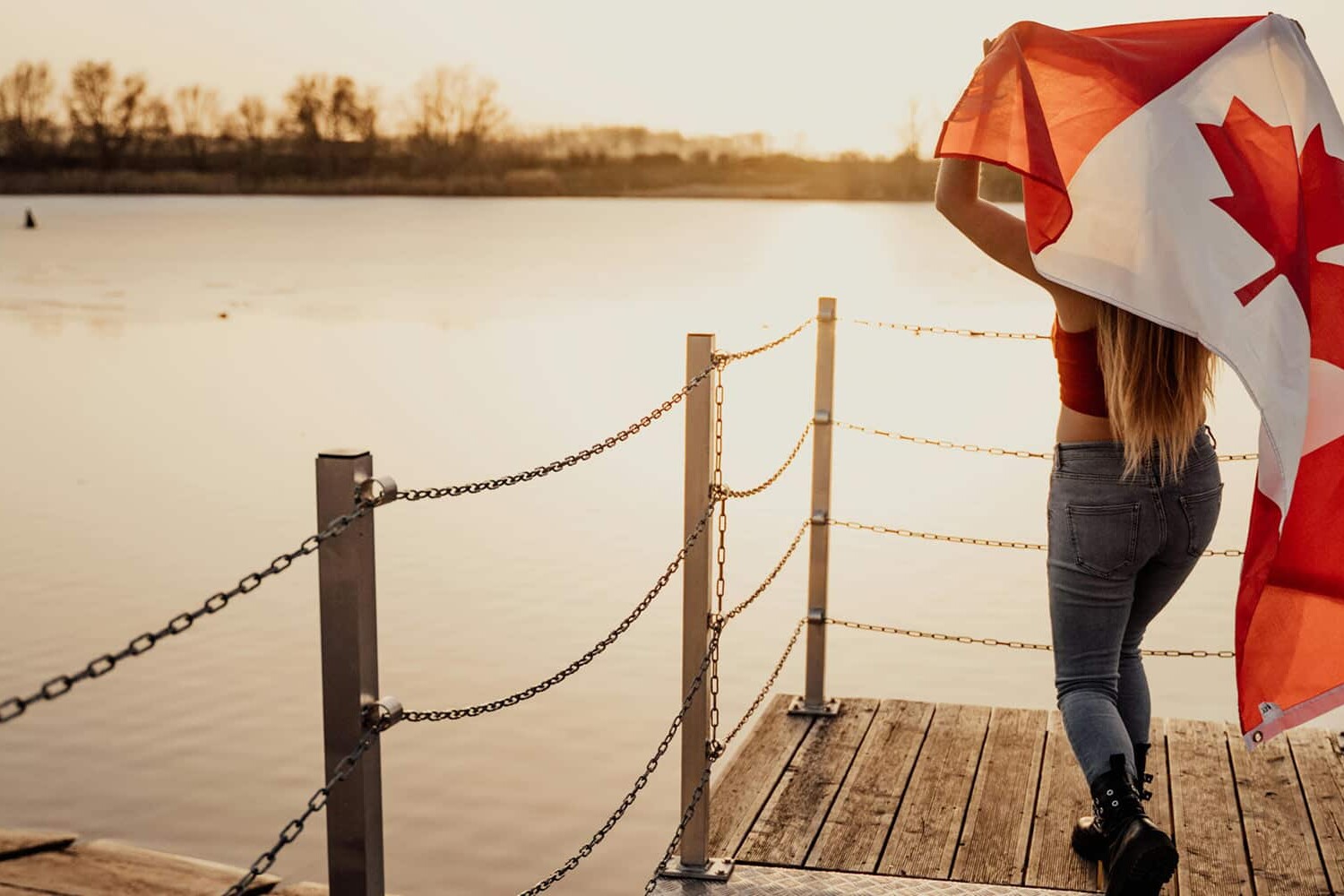 A person stands on a wooden dock by a lake at sunset, holding and partially wrapped in a large Canadian flag—a scene that beautifully reflects the Canadian values that were the deciding factor in this election.