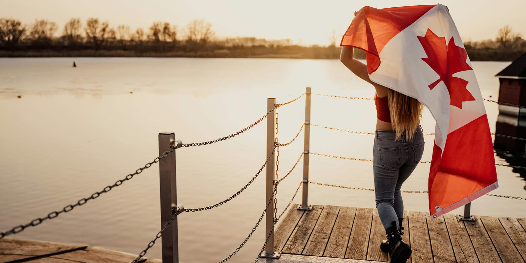 A person stands on a wooden dock by a lake at sunset, holding and partially wrapped in a large Canadian flag—a scene that beautifully reflects the Canadian values that were the deciding factor in this election.