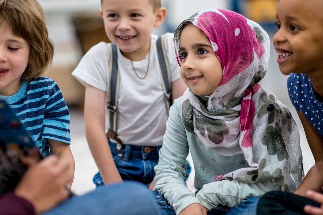A group of young children sit together on the floor, smiling and attentively listening to an adult read 