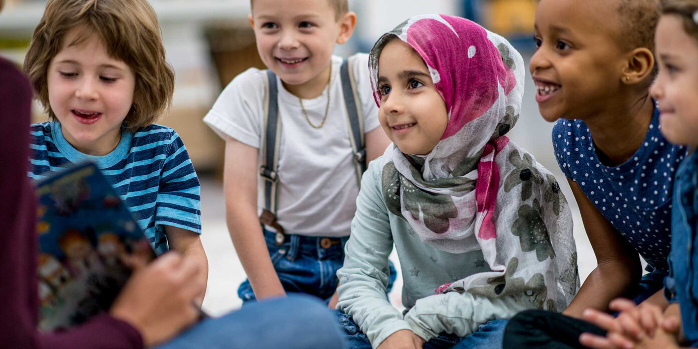 A group of young children sit together on the floor, smiling and attentively listening to an adult read 