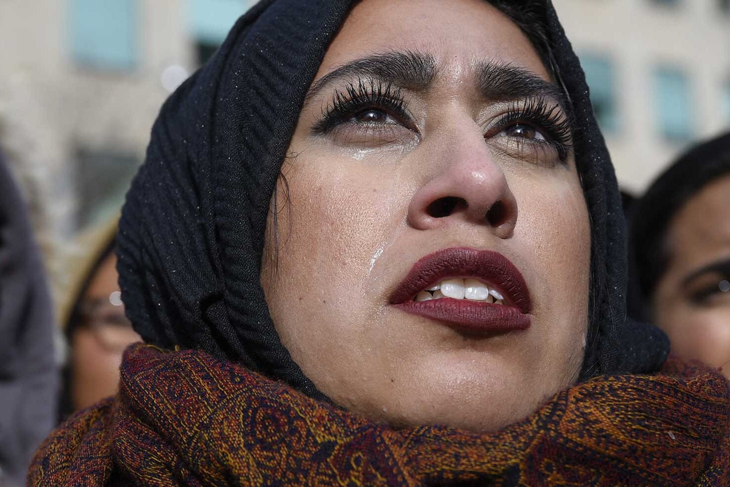 Three women stand close together outdoors. The woman in the center, wearing a headscarf and patterned scarf, looks upward with tears on her cheek—capturing the emotions highlighted in the Global Survey on Women as National Leaders.