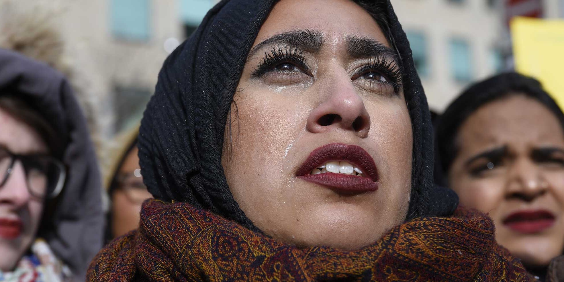 Three women stand close together outdoors. The woman in the center, wearing a headscarf and patterned scarf, looks upward with tears on her cheek—capturing the emotions highlighted in the Global Survey on Women as National Leaders.