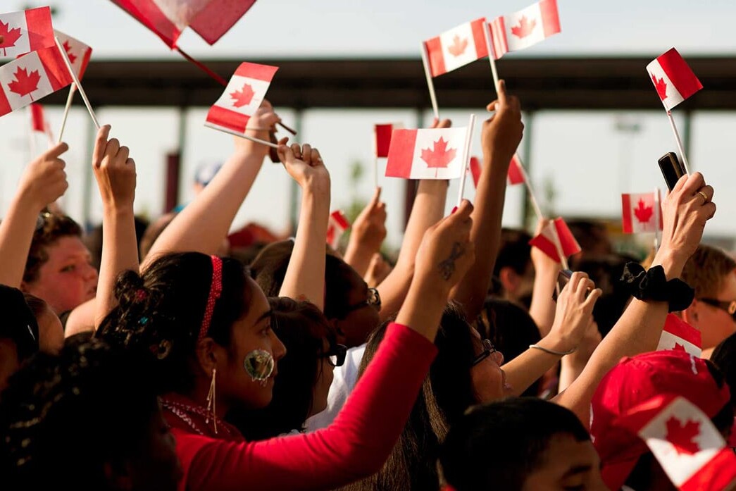 A crowd of people cheer and wave small Canadian flags, celebrating outdoors on a sunny day. Many wear red and white, showing national pride—proof that it's not happening here; Canadians keep faith in their democracy and government.