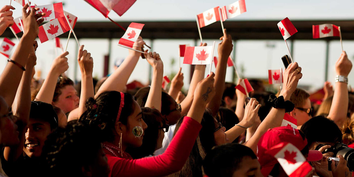 A crowd of people cheer and wave small Canadian flags, celebrating outdoors on a sunny day. Many wear red and white, showing national pride—proof that it's not happening here; Canadians keep faith in their democracy and government.