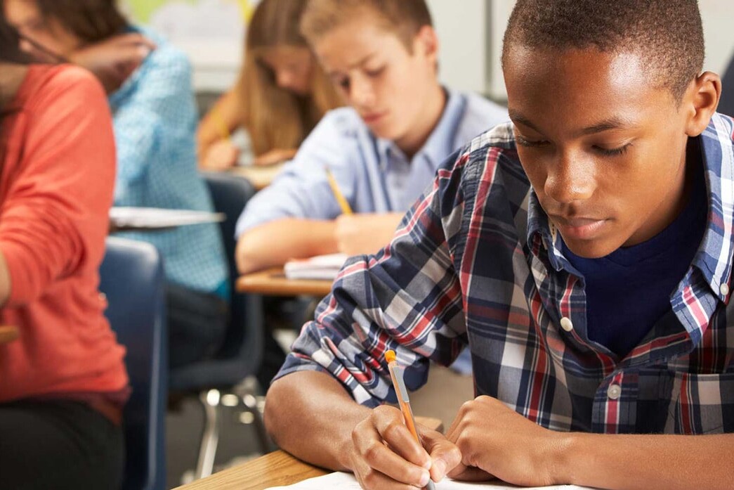 A group of students sitting at desks in a classroom, focused on writing or taking a test. As they work individually on their assignments, let’s not dismiss the painful pattern of microaggressions that can shape their experiences.