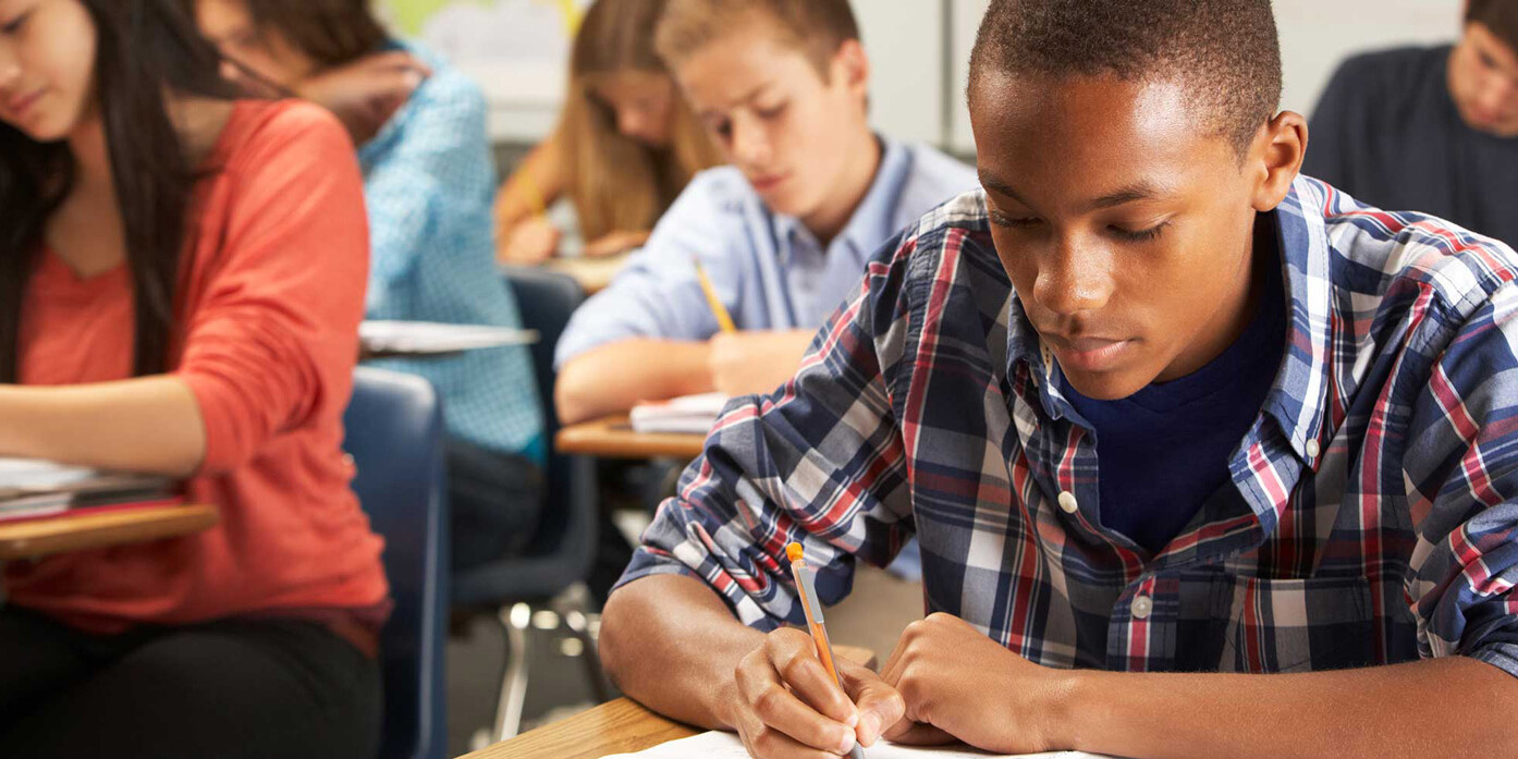 A group of students sitting at desks in a classroom, focused on writing or taking a test. As they work individually on their assignments, let’s not dismiss the painful pattern of microaggressions that can shape their experiences.