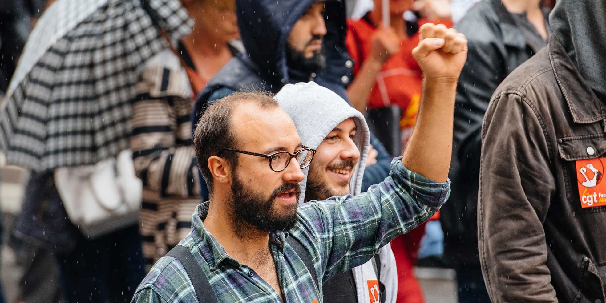 A group of people in casual clothing march in the rain. One man in glasses and a plaid shirt raises his fist, reflecting a sense of unity and purpose—reminding us that democracies are stronger than you may think.