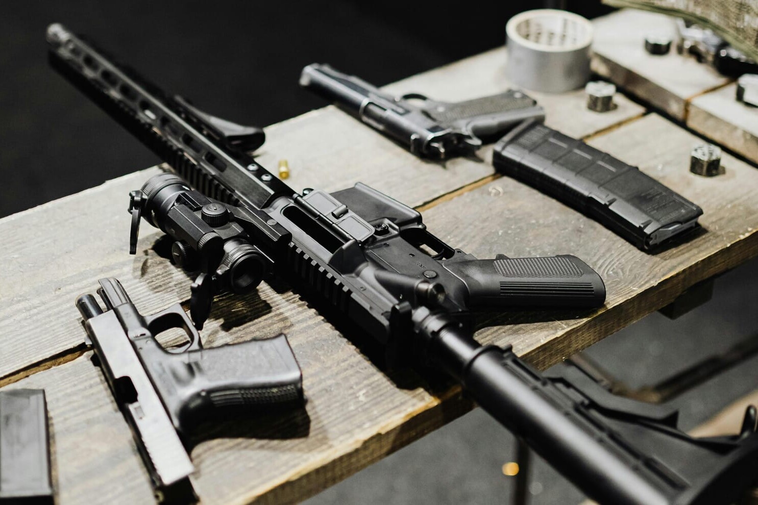 A wooden table displays several firearms, including a rifle and handguns, along with gun magazines, ammunition, and a roll of tape—a stark snapshot of why gun control is a lost cause in America.