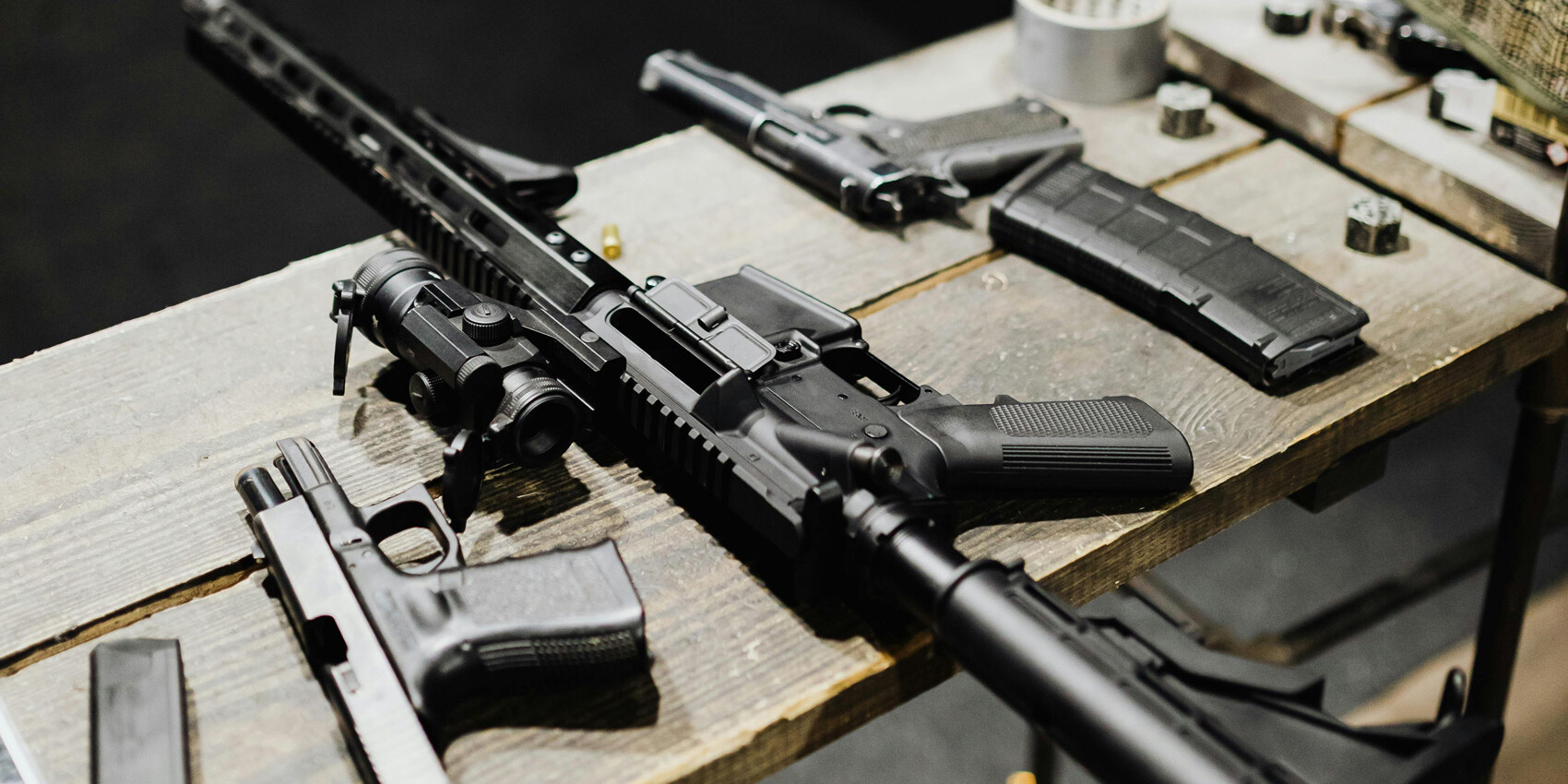 A wooden table displays several firearms, including a rifle and handguns, along with gun magazines, ammunition, and a roll of tape—a stark snapshot of why gun control is a lost cause in America.