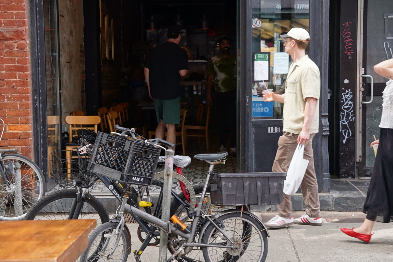 Two people walk past parked bikes outside a café or restaurant, perhaps discussing three ways that Toronto could become more autonomous from Ontario. Inside the dimly lit establishment, a few people can be seen enjoying their meals.