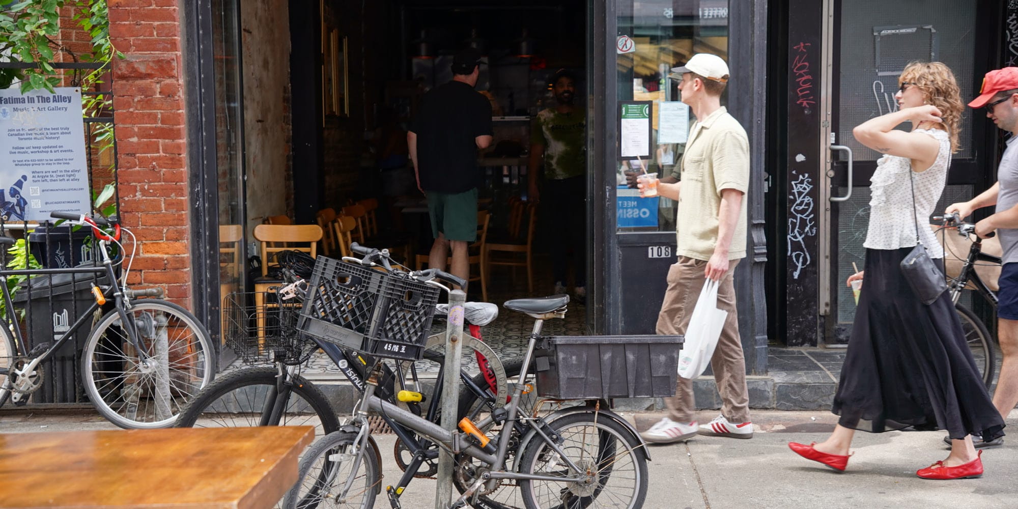 Two people walk past parked bikes outside a café or restaurant, perhaps discussing three ways that Toronto could become more autonomous from Ontario. Inside the dimly lit establishment, a few people can be seen enjoying their meals.