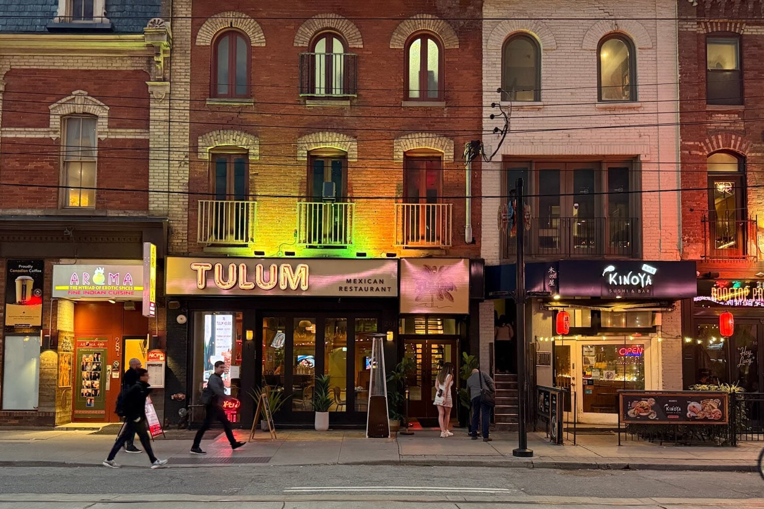 Street view of colorful brick buildings with various shops and restaurants, including Tulum Mexican Restaurant. As people walk on the sidewalk and a cyclist passes by in the evening, the scene reflects three ways that Toronto could become more autonomous from Ontario.