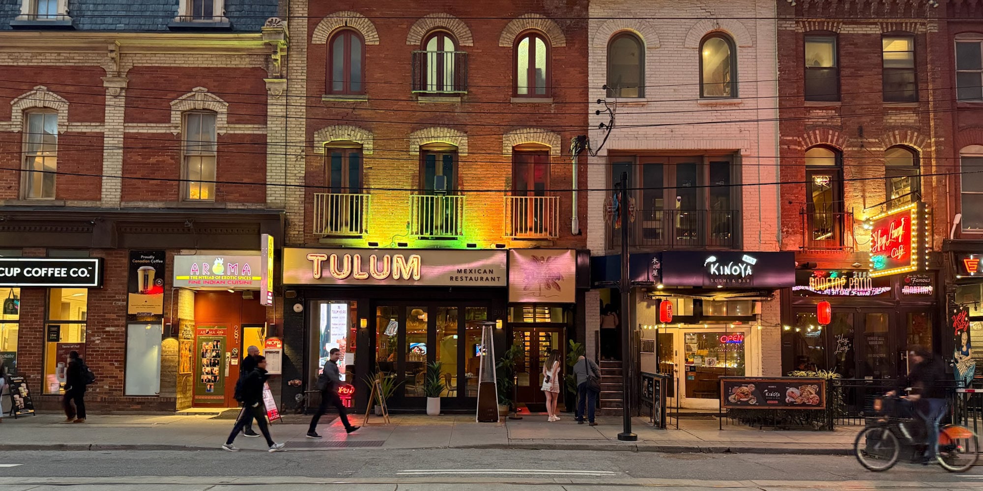 Street view of colorful brick buildings with various shops and restaurants, including Tulum Mexican Restaurant. As people walk on the sidewalk and a cyclist passes by in the evening, the scene reflects three ways that Toronto could become more autonomous from Ontario.