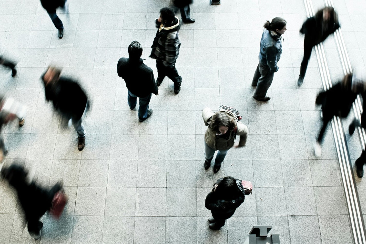 An overhead view of people walking and standing on a tiled floor in a public indoor space, some caught in motion blur. The scene prompts reflection: ahead of a federal election, what road will Conservatives take on immigration?.