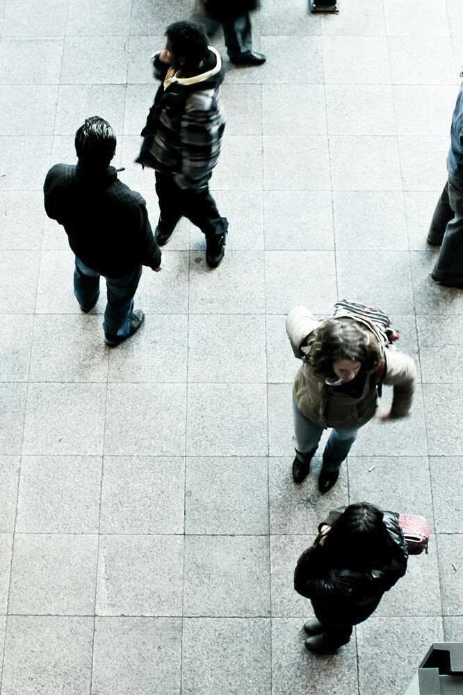 An overhead view of people walking and standing on a tiled floor in a public indoor space, some caught in motion blur. The scene prompts reflection: ahead of a federal election, what road will Conservatives take on immigration?.