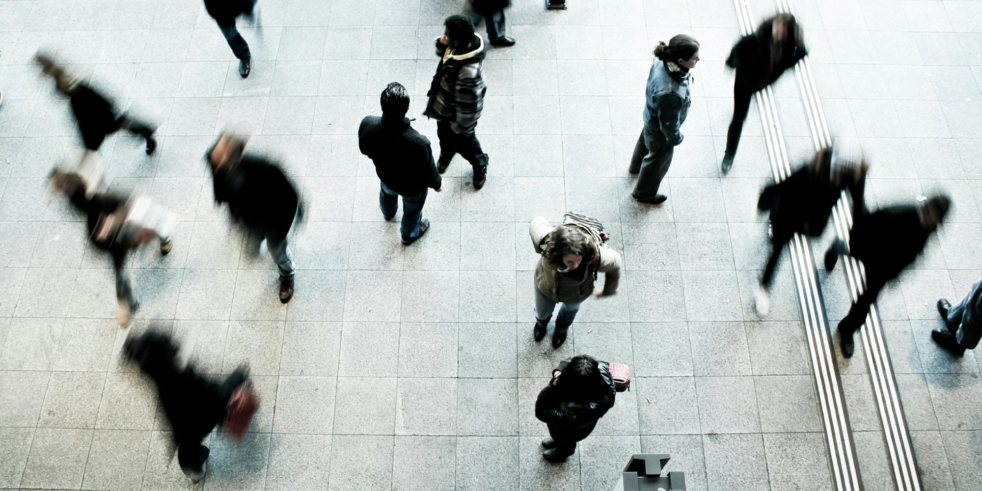An overhead view of people walking and standing on a tiled floor in a public indoor space, some caught in motion blur. The scene prompts reflection: ahead of a federal election, what road will Conservatives take on immigration?.