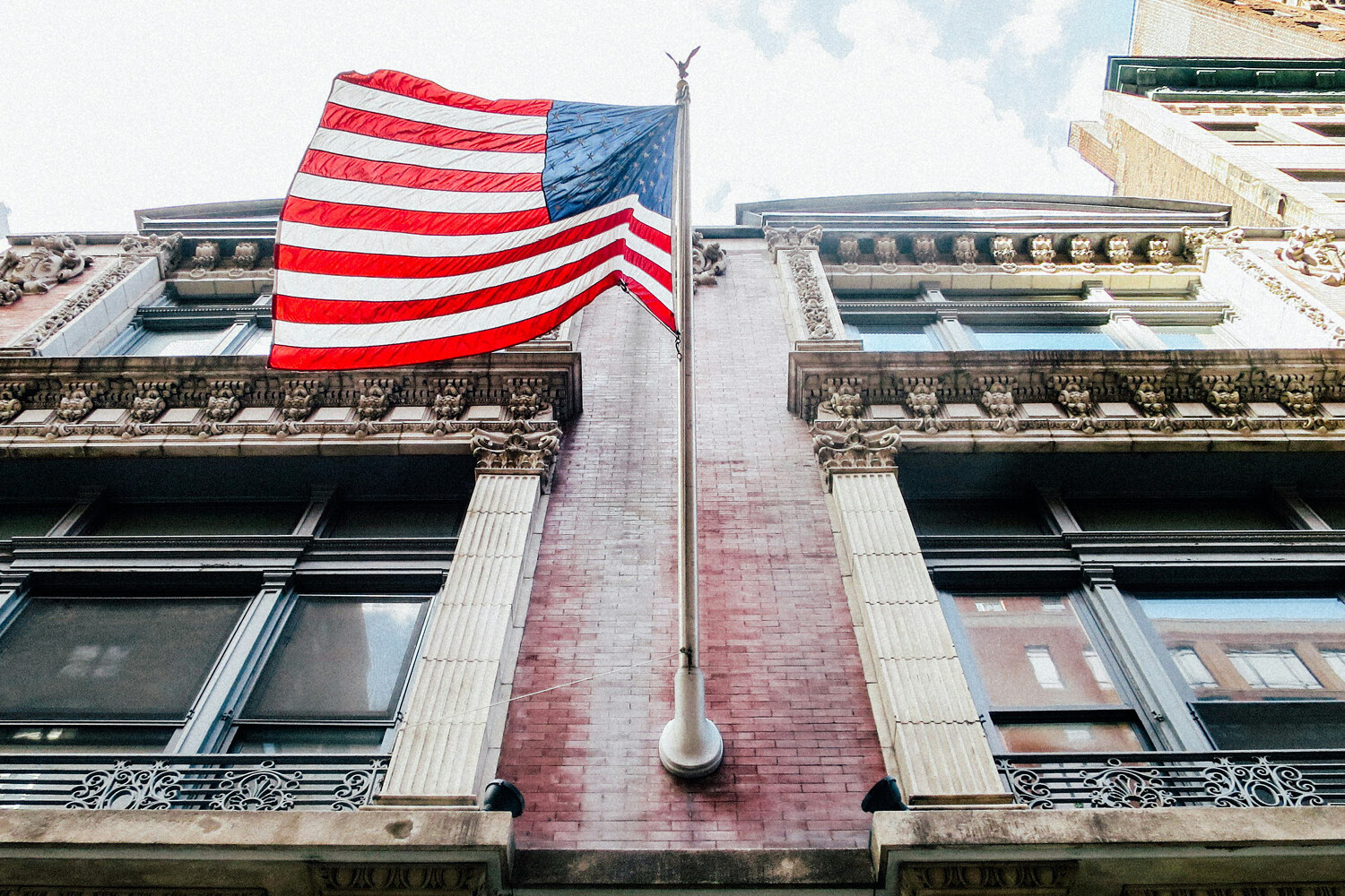 An American flag waves atop a flagpole on a historic brick building, its colors undimmed even as Canada’s opinion of the US sits at an unprecedented low, with ornate window frames and tall city buildings under a partly cloudy sky.