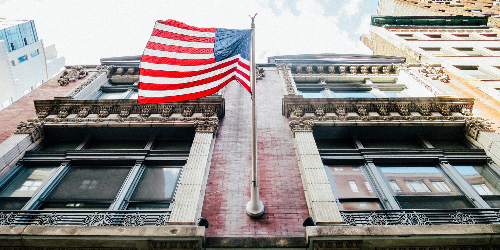 An American flag waves atop a flagpole on a historic brick building, its colors undimmed even as Canada’s opinion of the US sits at an unprecedented low, with ornate window frames and tall city buildings under a partly cloudy sky.