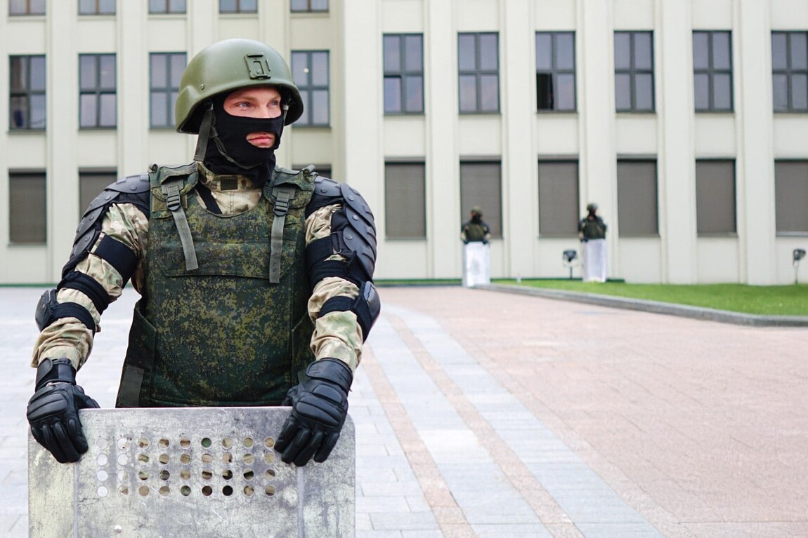 A soldier in camouflage gear and helmet holds a riot shield, standing guard in front of a government building—a scene that raises the question: The authoritarian reflex: Will it manifest in Canada? Two other armed guards are visible near the windows.