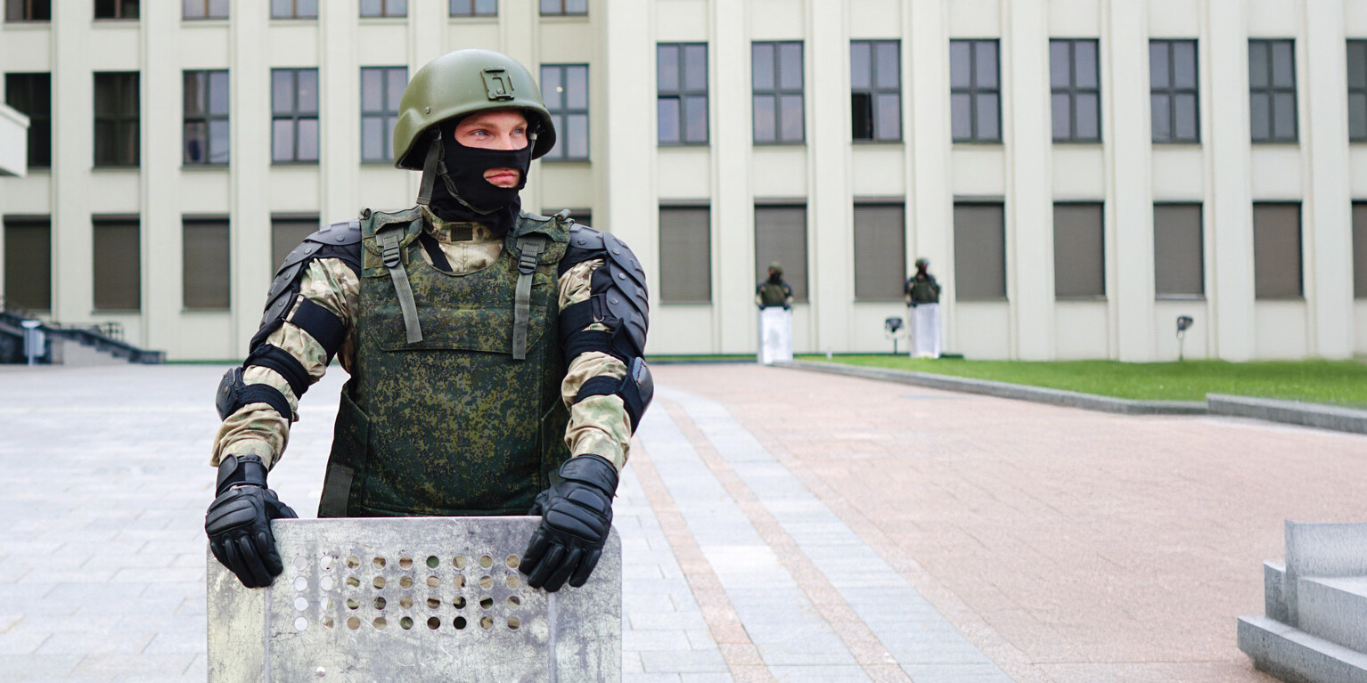 A soldier in camouflage gear and helmet holds a riot shield, standing guard in front of a government building—a scene that raises the question: The authoritarian reflex: Will it manifest in Canada? Two other armed guards are visible near the windows.