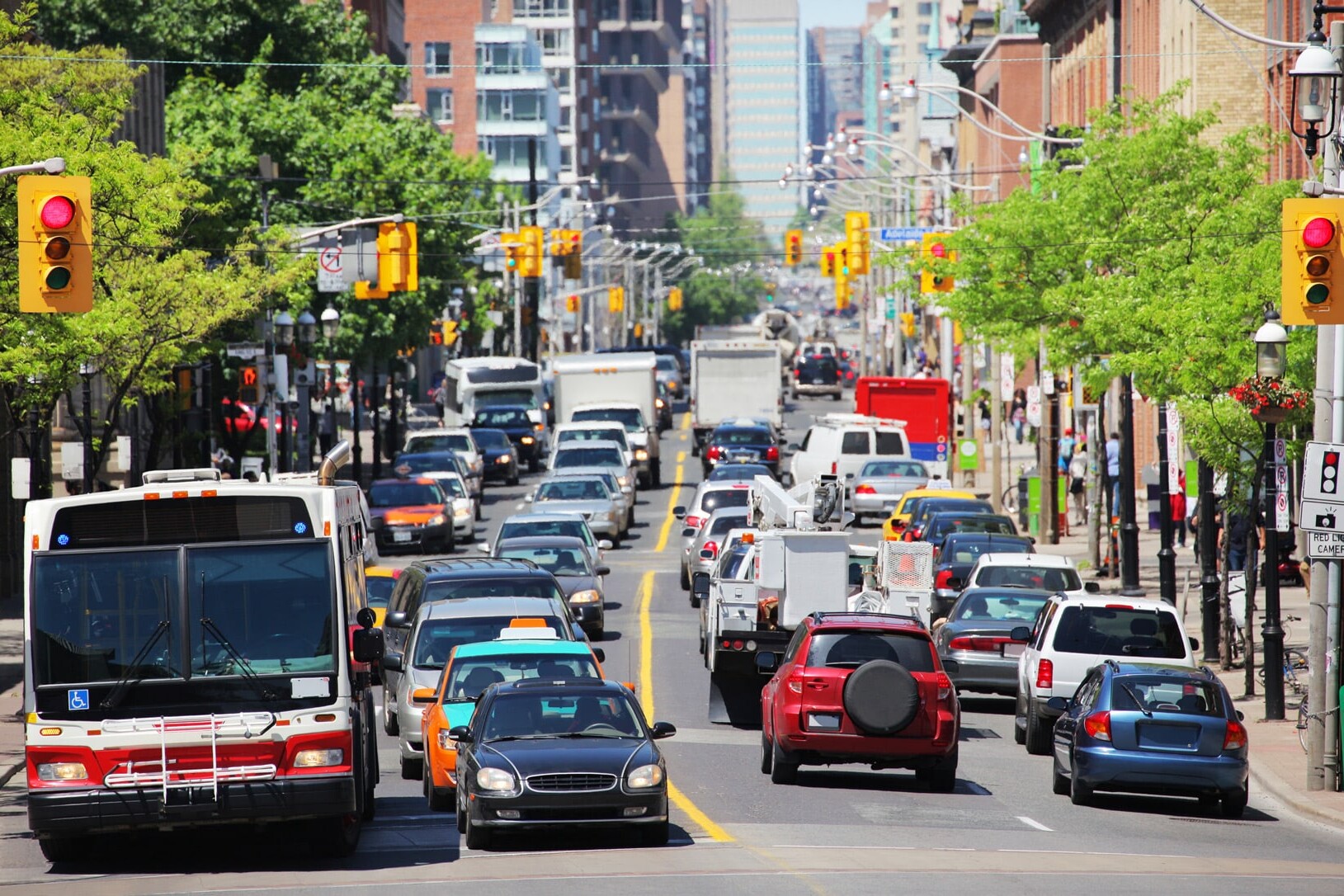 A busy city street filled with cars, trucks, taxis, and a public bus—reflecting Canadians’ Satisfaction with Public Services—waits at a traffic light, surrounded by buildings and trees on a sunny day.