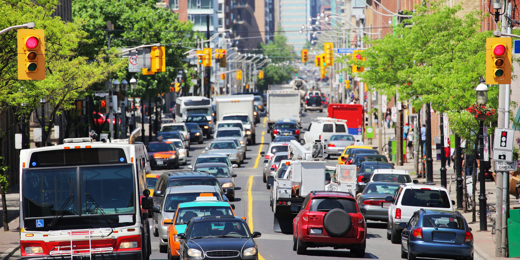 A busy city street filled with cars, trucks, taxis, and a public bus—reflecting Canadians’ Satisfaction with Public Services—waits at a traffic light, surrounded by buildings and trees on a sunny day.
