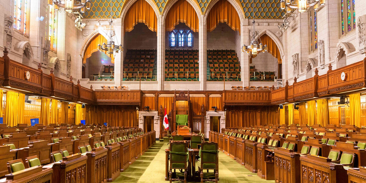 The interior of a grand parliamentary chamber with rows of green seats, ornate wood paneling, high arched ceilings, chandeliers, and a Canadian flag near the central speaker’s chair—an impressive setting for shaping perceptions of gender and political leadership.