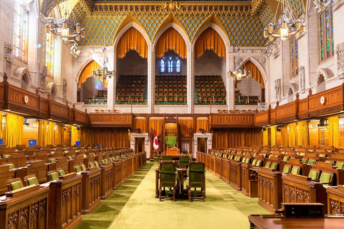 The interior of a grand parliamentary chamber with rows of green seats, ornate wood paneling, high arched ceilings, chandeliers, and a Canadian flag near the central speaker’s chair—an impressive setting for shaping perceptions of gender and political leadership.