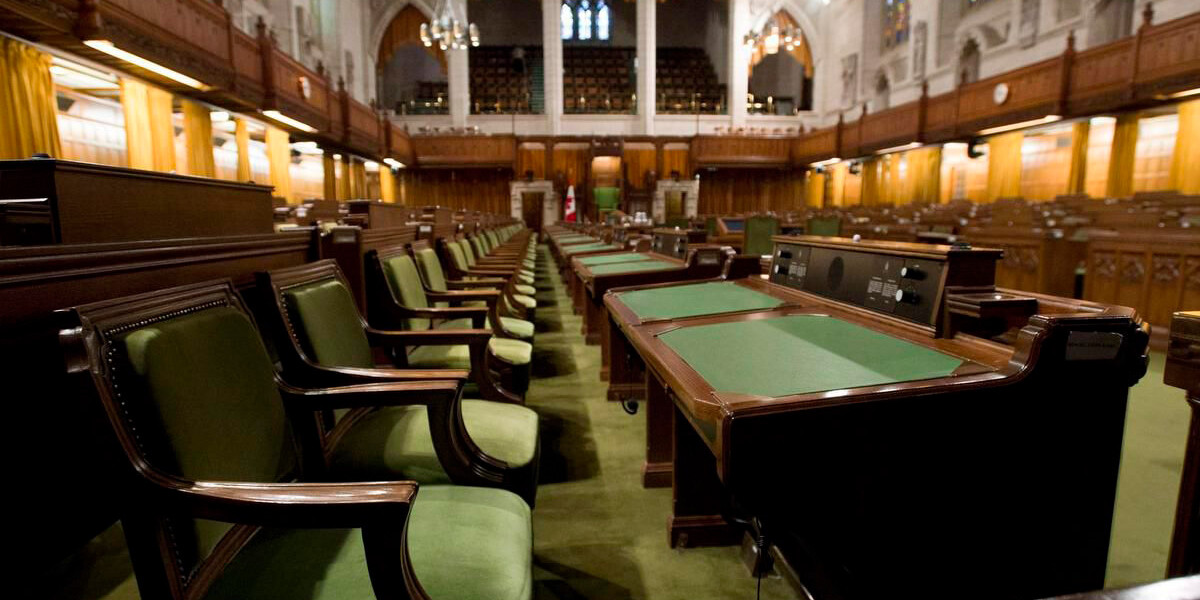 Rows of green-cushioned desks and chairs fill a grand parliamentary chamber with high arched ceilings, wood paneling, hanging lights, and stained-glass windows—reminding voters to be suspicious of all the magical promises from politicians.
