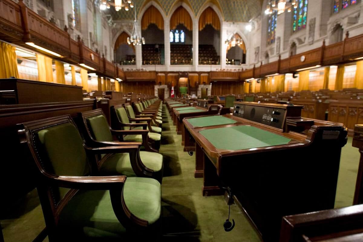Rows of green-cushioned desks and chairs fill a grand parliamentary chamber with high arched ceilings, wood paneling, hanging lights, and stained-glass windows—reminding voters to be suspicious of all the magical promises from politicians.